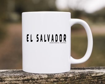 Puede incluir: Una taza de cerámica blanca con el texto negro EL SALVADOR impreso en una fuente negrita y estilizada, con el texto más pequeño MISAMIS ORIENTAL, PHILIPPINES situado directamente debajo de la inscripción principal en el frente.