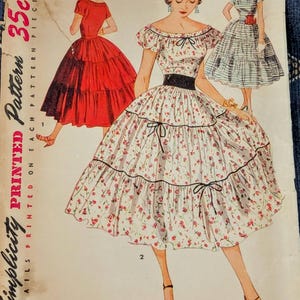 Op de afbeelding: Een Simplicity 4640 naaipatroon-envelop met illustraties van drie jurken. De middelste jurk is wit met een kleine rode bloemenprint, een zwarte tailleband en een gelaagde rok. De andere twee jurken zijn effen rood en blauw gestreept.