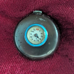 May include: A round, dark gray metal pocket watch featuring a white circular dial with black numerals, a red number 12, and black hands. The watch face is surrounded by a bright blue ridged bezel, and a small silver winding crown sits at the top.