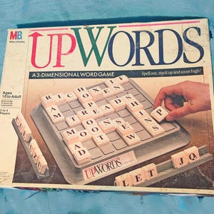 May include: Vintage Upwords board game box with the game board and tiles visible. The box features the game's title in red, green, and blue. The game board has tiles with letters on them. The box is designed for 2 to 4 players, ages 10 to adult.