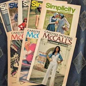 May include: A collection of vintage Simplicity and McCall's fashion news pattern catalogs. The covers feature various photographs of people in clothing, including a white vest and pants set, colorful balloons, and outdoor scenes with printed text.