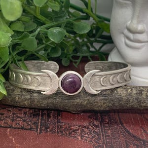 May include: Silver bracelet with a triple moon design and a purple gemstone in the center.