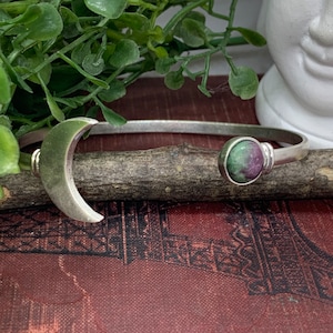 May include: A silver bracelet with a crescent moon charm and a green and pink gemstone. The bracelet is on a brown surface.