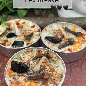 May include: Three small, round candles with the text "Hex Breaker" on a white background. The candles are silver and filled with a white wax base, herbs, spices, and a black stone. The candles have a decorative theme.