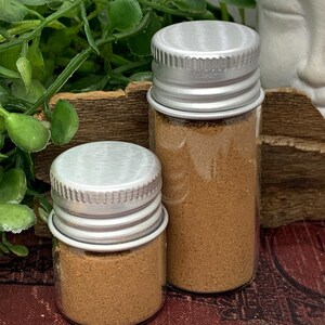 May include: Two small glass jars with silver lids, filled with a brown powder. The jars are different sizes and sit on a dark red surface. Green foliage and a white statue are in the background.