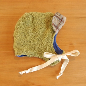 May include: A green fuzzy hat with a plaid fabric ear flap and a button. The hat has a blue lining and a white ribbon tie.