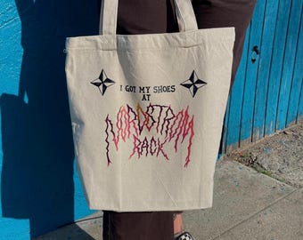 Puede incluir: Bolso tote color crema con el texto "I GOT MY SHOES AT NORDSTROM RACK" en negro y gráficos de estrellas. El bolso tiene forma rectangular con dos asas. La persona lleva zapatos a cuadros blancos y negros.