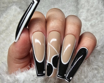 Pode incluir: Uma mão exibindo unhas artificiais longas e quadradas. As unhas apresentam uma base bege com desenhos em preto e branco. Algumas unhas têm uma borda preta com detalhes brancos, outras arte de linha branca abstrata.