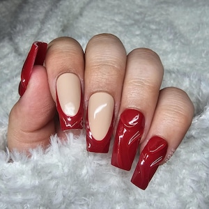 May include: A hand showing long, square-shaped artificial nails. The nails are a glossy, deep red colour with a French tip design on some, while others are solid red with a textured pattern. The nails are set against a soft, white background.