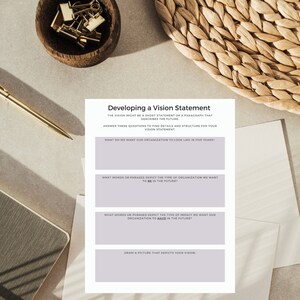 May include: A vision statement worksheet with prompts to define organizational goals. The worksheet is on a white paper with a light purple border. A gold pen, a small wooden bowl with gold binder clips, and a woven placemat are also in the image.