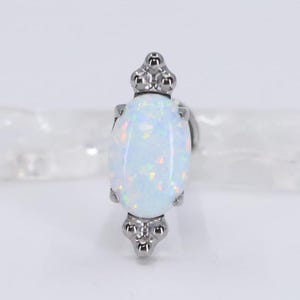 May include: A silver-toned earring with an oval, opalescent gemstone. The gemstone has a pearly white base with flashes of color. The earring has decorative silver accents at the top and bottom, giving it a vintage or antique look.