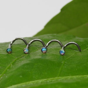 Puede incluir: Cuatro anillos de nariz plateados con piedras preciosas de ópalo en diferentes tonos de azul, verde y rosa. Los anillos están dispuestos en una fila sobre una hoja verde.