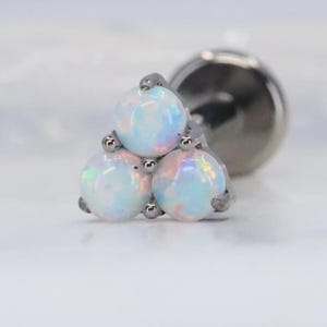 Può includere: Primo piano di un orecchino color argento con tre pietre di opale bianche disposte in un gruppo triangolare. Gli opali hanno una lucentezza perlescente, che riflette sfumature di blu e rosa. Il perno e il retro dell'orecchino sono anch'essi argentati.