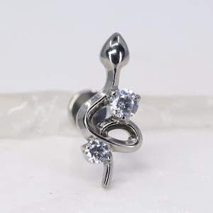May include: A silver-coloured stud earring shaped like a snake. The snake design incorporates two clear, round gemstones. The earring has a post backing. The snake's head is at the top of the design.