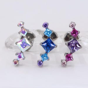 May include: Three silver stud earrings, each featuring a cluster of colorful, square and round gemstones. The earrings display a gradient of colors, including iridescent, blue, purple, and pink stones. The earrings are designed for pierced ears.