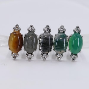 Könnte beinhalten: Fünf Edelsteinringe mit ovalen Steinen in verschiedenen Farben: Bernstein, Grau, Schwarz, Grün und Jade. Jeder Ring hat eine silberfarbene dekorative Fassung.