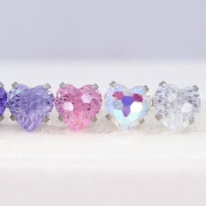 May include: A row of four heart-shaped stud earrings featuring faceted stones in purple, pink, iridescent, and clear colours. Each stone is secured by four small silver-toned metal prongs, creating a uniform display of sparkling jewellery pieces.