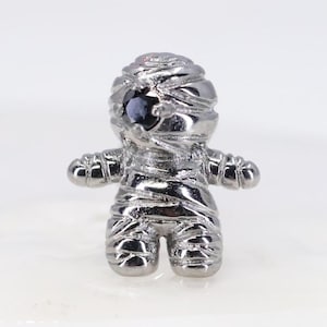 Pode incluir: Um berloque de metal prateado em forma de múmia, com uma superfície texturizada que imita faixas enroladas. A figura possui uma única pedra preciosa redonda e escura incrustada na cabeça como um olho, com um acabamento polido e brilhante.