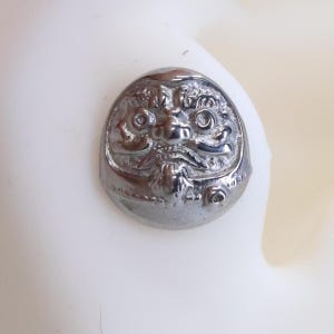 Può includere: Un ciondolo a forma di bambola Daruma color argento con dettagliati tratti del viso, tra cui grandi occhi, un naso e dei baffi. Il ciondolo è rotondo e ha una parte superiore leggermente a cupola, che gli conferisce un aspetto tridimensionale. Lo sfondo è di colore bianco sporco.