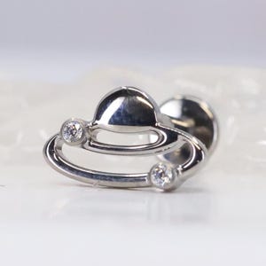 May include: A silver-colored, planet-shaped plug with two small, clear gemstones. The plug has a rounded top and a curved, ring-like design with a gemstone on each end. The background is a soft, blurred white.