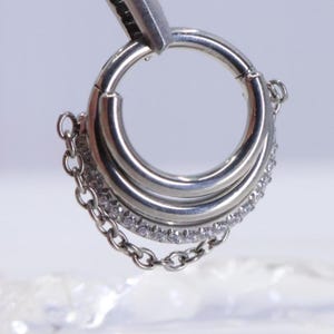 May include: A silver-colored hinged hoop earring with three stacked rings. The bottom ring is encrusted with small, clear gemstones. A delicate chain dangles from the earring, adding a touch of elegance. The earring is held by a silver tool.