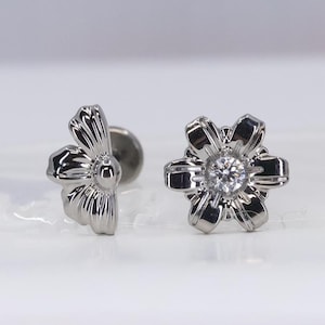 Puede incluir: Un par de pendientes de botón plateados con forma de flor. Un pendiente presenta una gema central, mientras que el otro muestra los pétalos de la flor. Los pendientes están sobre un fondo blanco.