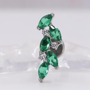 May include: A silver-toned earring featuring a cluster of marquise-cut green gemstones and small, round, clear gemstones. The design resembles a stylized leaf or floral arrangement, with the gems set in a curved, elegant pattern.