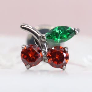 May include: A silver-toned metal stud earring shaped like a pair of cherries. The design features two round, faceted red stones representing the fruit and one marquise-cut green stone representing a leaf, all set in a polished metallic frame.