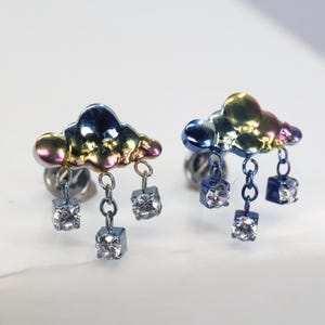 May include: A pair of rainbow-colored cloud earrings with dangling cubic zirconia stones. Each earring features a cloud-shaped charm with a rainbow iridescent finish and three dangling square-shaped cubic zirconia stones.