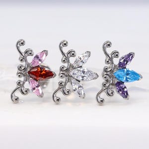Puede incluir: Tres pendientes plateados con forma de flor y detalles de piedras preciosas de colores. Un pendiente presenta piedras rojas y rosas, otro tiene piedras transparentes y el último tiene piedras azules y moradas. Los pendientes tienen un diseño decorativo y ondulado.