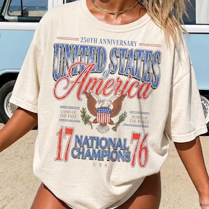 Könnte beinhalten: Ein elfenbeinfarbenes Kurzarm-T-Shirt mit einem Grafikdruck mit dem Text 250TH ANNIVERSARY UNITED STATES OF AMERICA, LAND OF THE FREE, HOME OF THE BRAVE, 17 NATIONAL CHAMPIONS 76 und einem zentralen Adler-Emblem mit einem Schild.