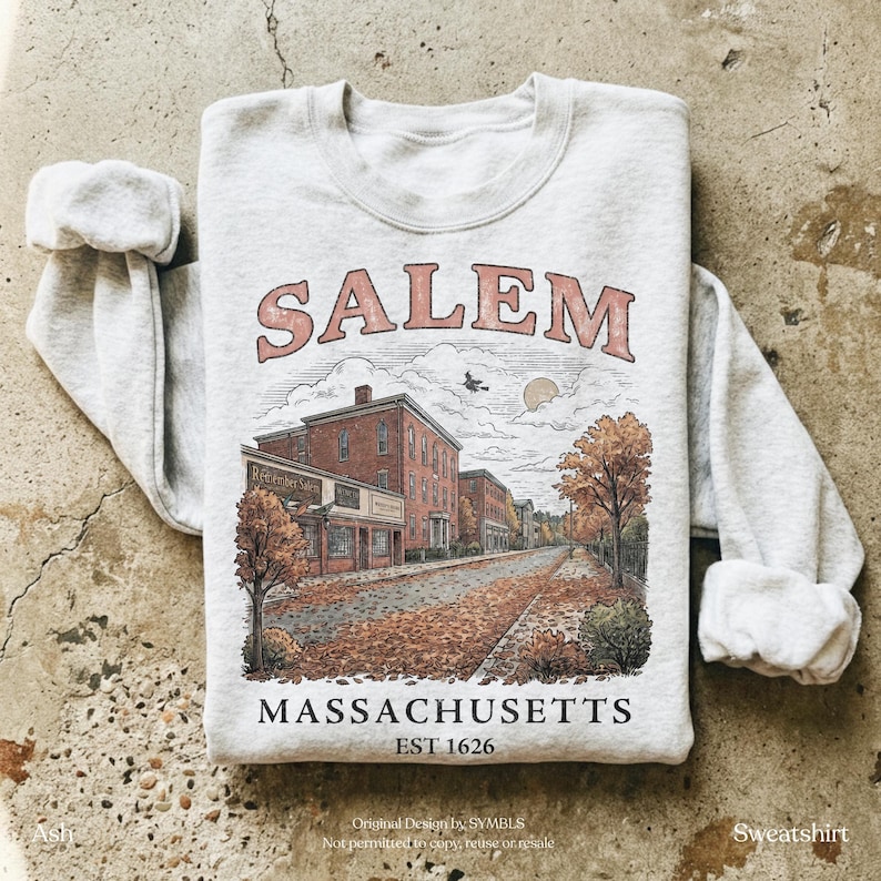 K&ouml;nnte beinhalten: Ein aschgraues Sweatshirt mit Rundhalsausschnitt und einem Grafikdruck einer Stra&szlig;enszene in Salem, Massachusetts. Das Design zeigt den Text SALEM in geschwungenen Buchstaben, MASSACHUSETTS und EST 1626 sowie Backsteingeb&auml;ude und B&auml;ume.