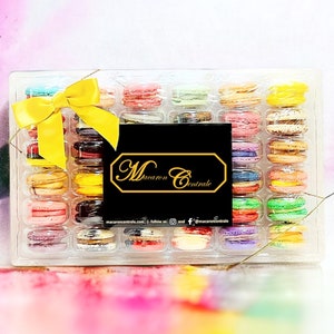 May include: A clear plastic box filled with colorful macarons. The box is tied with a yellow ribbon and has a black label with the text "Macaron Centrale" on it.
