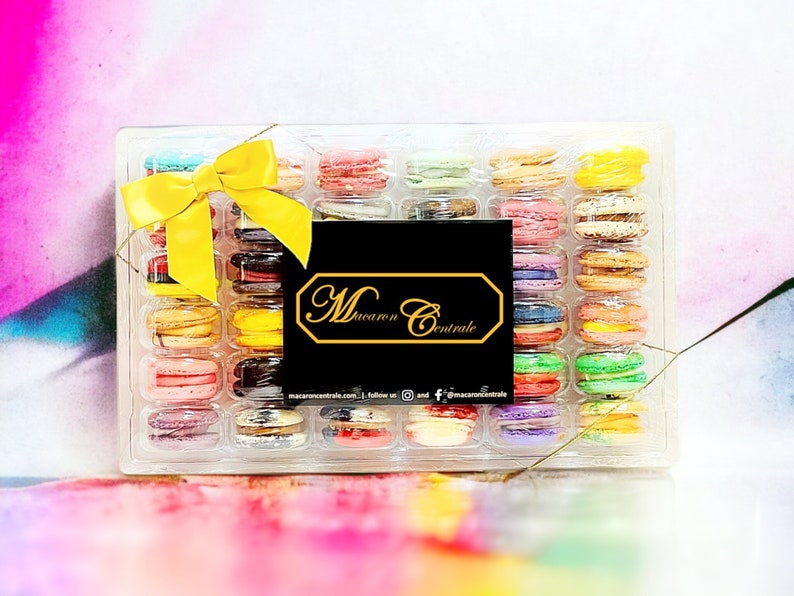 May include: A clear plastic box filled with colorful macarons. The box is tied with a yellow ribbon and has a black label with the text "Macaron Centrale" on it.
