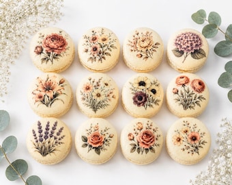 May include: Twelve round macarons with floral designs. Each macaron features a different flower, including roses, lilies, and hydrangeas, in shades of pink, peach, and purple. The macarons are arranged on a white surface.