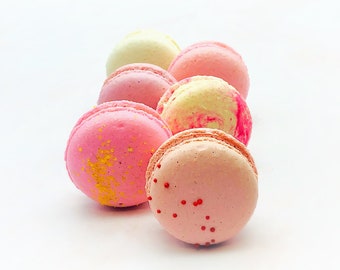 May include: A close-up of six pastel-colored macarons. The macarons are arranged in a row, with the bottom macaron having red sprinkles. The macarons are on a white surface.