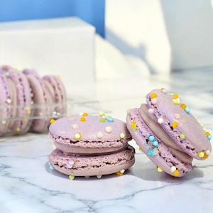 May include: Two purple macarons with colorful sprinkles, stacked on top of each other. The macarons are on a white marble surface.