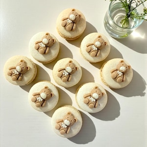 May include: A collection of ten macarons, each topped with a small, brown teddy bear decoration. The macarons have a light beige color and are arranged in a circular pattern on a white surface. A small glass vase with white flowers is in the background.