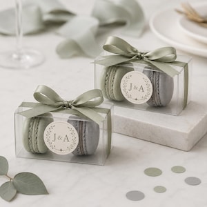May include: Two clear boxes each contain two macarons, tied with sage green ribbon bows. Each macaron box has a circular sticker with the initials "J & A" inside a wreath. The macarons are in shades of green and gray.