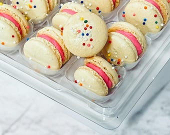 May include: A clear plastic container filled with colorful macarons. The macarons are white with pink filling and decorated with colorful sprinkles.