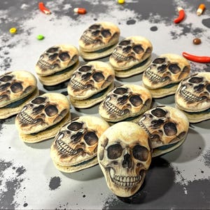 May include: An overhead shot displays a collection of skull-shaped cookies, each with detailed facial features. The cookies are light-colored with darker shading, arranged on a gray surface with scattered candies.