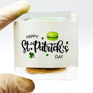 K&ouml;nnte beinhalten: Ein durchsichtiger Plastikbeh&auml;lter mit einem wei&szlig;en Etikett, auf dem "Happy St. Patrick's Day" steht und ein gr&uuml;nes Macaron abgebildet ist. Der Beh&auml;lter ist mit einer hellbraunen Substanz gef&uuml;llt.