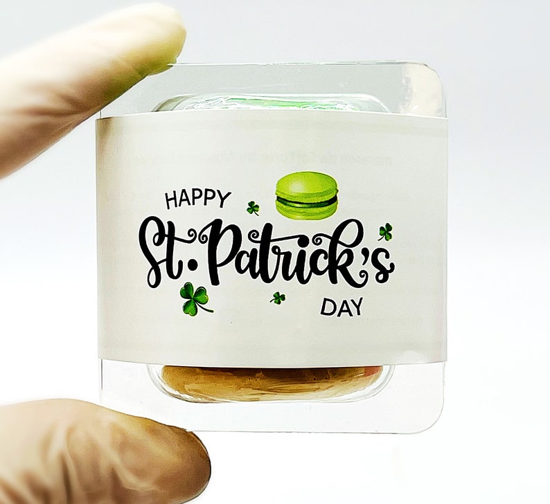K&ouml;nnte beinhalten: Ein durchsichtiger Plastikbeh&auml;lter mit einem wei&szlig;en Etikett, auf dem "Happy St. Patrick's Day" steht und ein gr&uuml;nes Macaron abgebildet ist. Der Beh&auml;lter ist mit einer hellbraunen Substanz gef&uuml;llt.