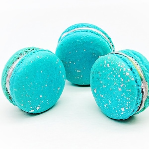 May include: Three turquoise macarons with silver accents and a white background. The macarons have a speckled appearance and a light-colored filling. The image is a close-up of the desserts.