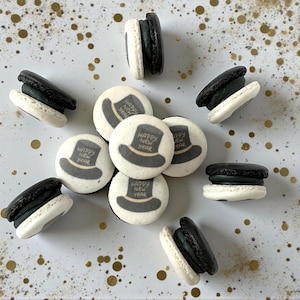 May include: An assortment of macarons, some black and white, others with a white base and a top hat design that reads "Happy New Year". The macarons are scattered on a white surface with gold confetti.