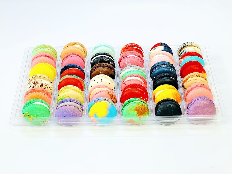 May include: A clear plastic tray filled with colorful macarons. The macarons are arranged in rows and are a variety of colors, including pink, blue, green, yellow, and purple.