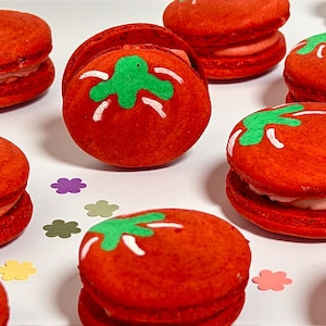 May include: A collection of red macarons decorated to resemble tomatoes. Each macaron has a green stem and white accents. The macarons are arranged on a white surface with colorful flower-shaped confetti.