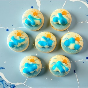 May include: Seven round cookies decorated with a blue and white cloud design, a yellow sun, and blue wave details. The cookies are arranged on a light blue surface with a marbled effect.