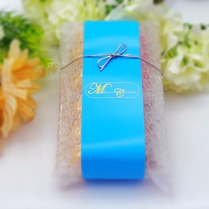 May include: A blue and white gift box with a white ribbon and bow. The box is wrapped in clear plastic with bubble wrap. The box has the text "M. Sweets" on the front.