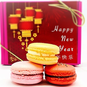 May include: A pink box with a red background and gold Chinese lanterns. The box says "Happy New Year" and "新年快乐" in Chinese. Three macarons are in front of the box: one yellow, one pink, and one red.